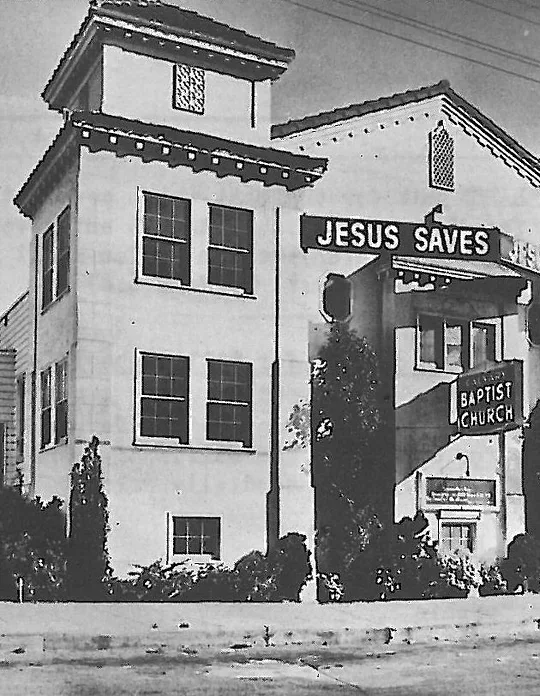 A historic black-and-white photograph of Calvary Baptist Church, a two-story mission-style building with a rooftop JESUS SAVES sign