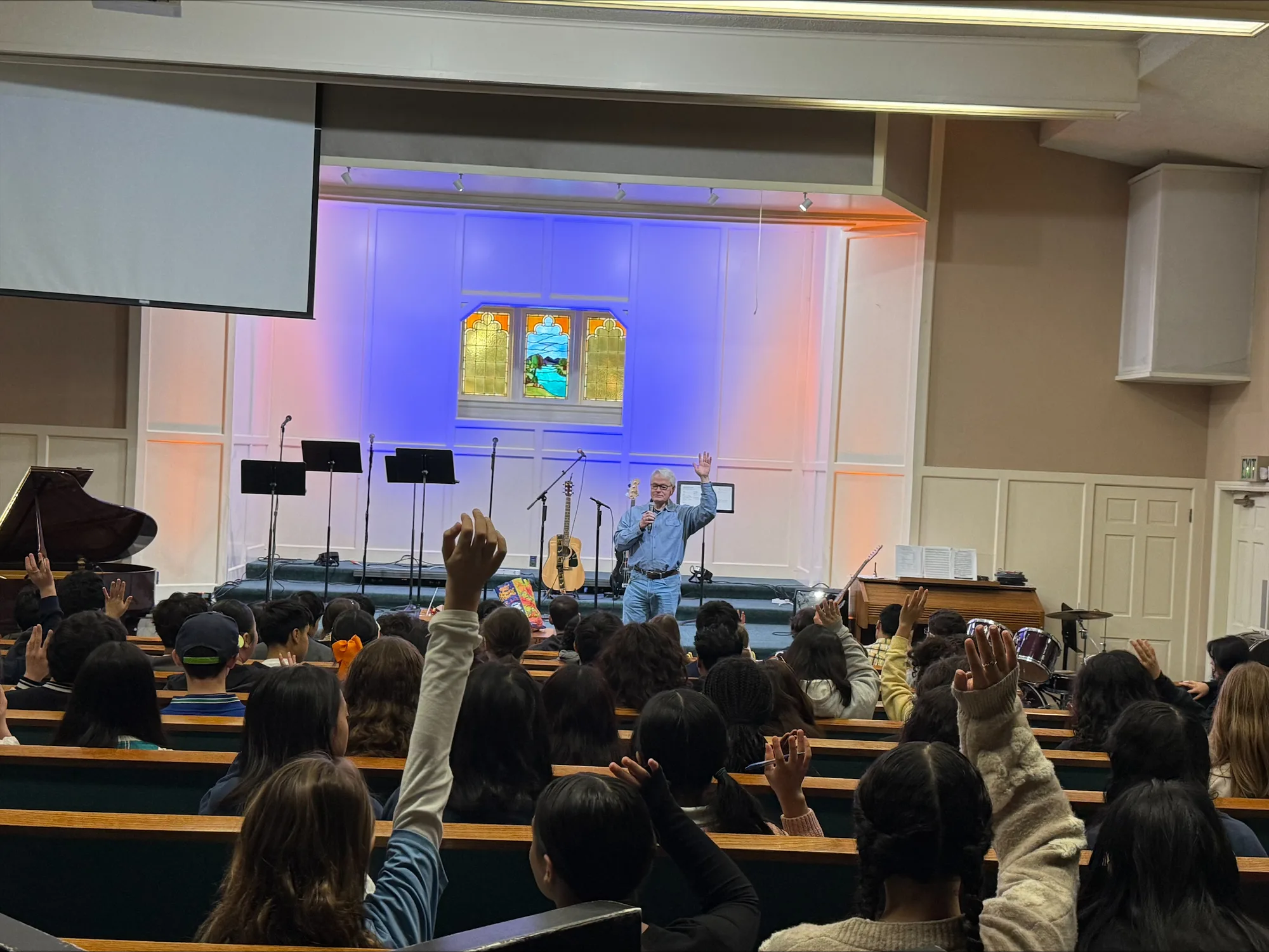 Students with hands raised in the pews during a chapel message, the speaker on stage below the sanctuary's stained-glass window