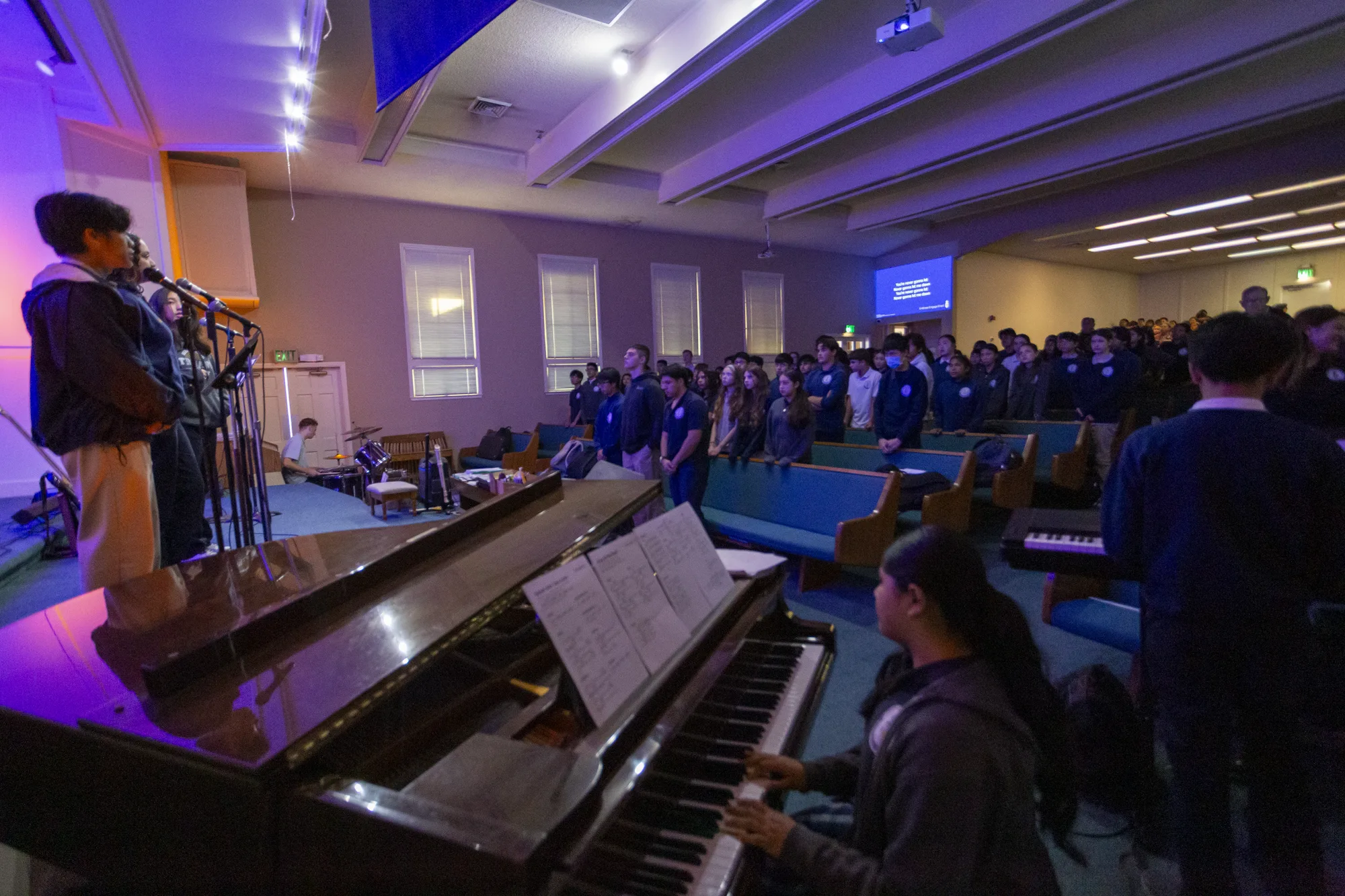 The whole student body standing in worship during chapel, a student trio at the microphones and a student at the piano