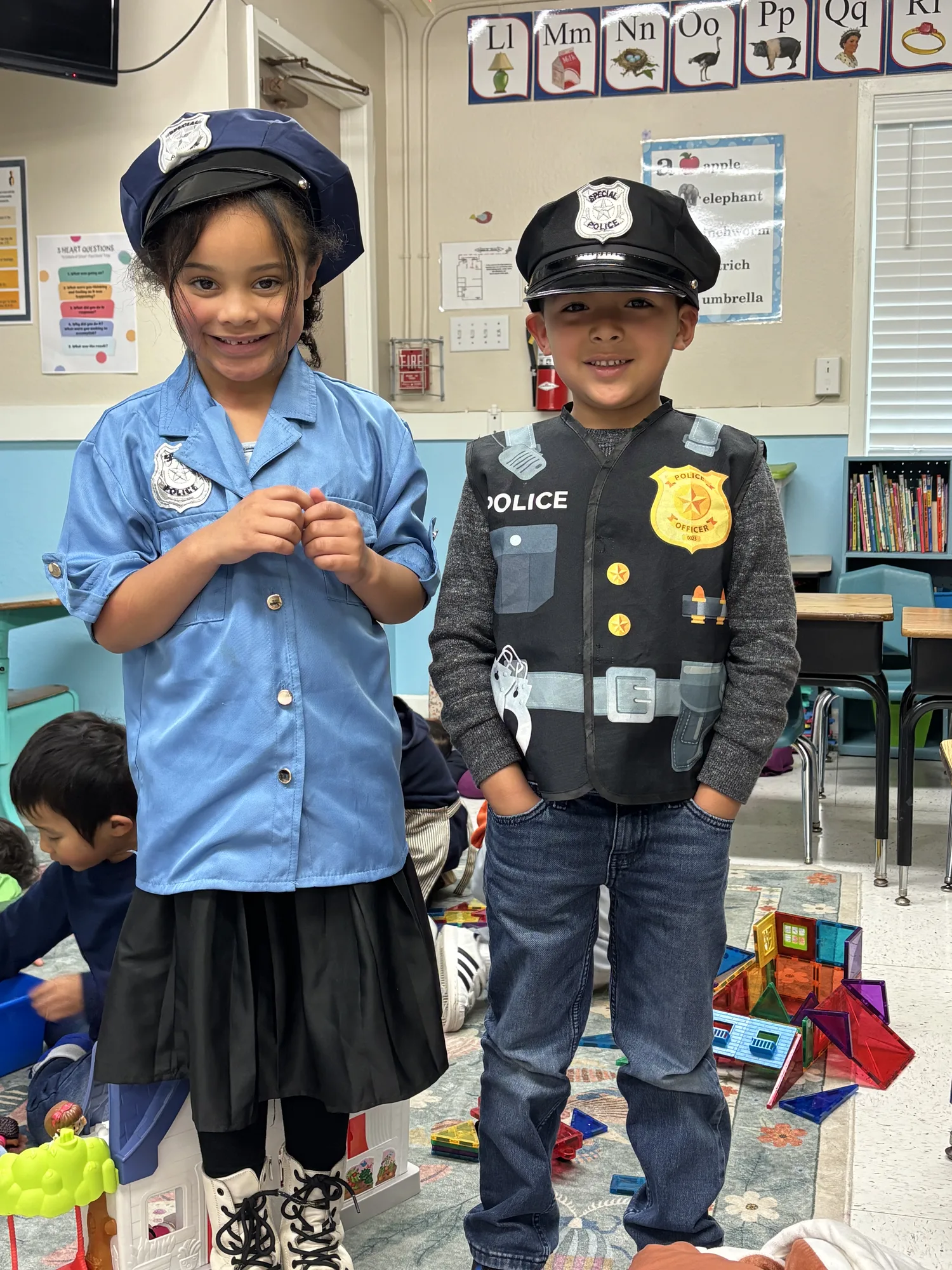 Two kindergartners in police dress-up costumes smiling by the alphabet wall