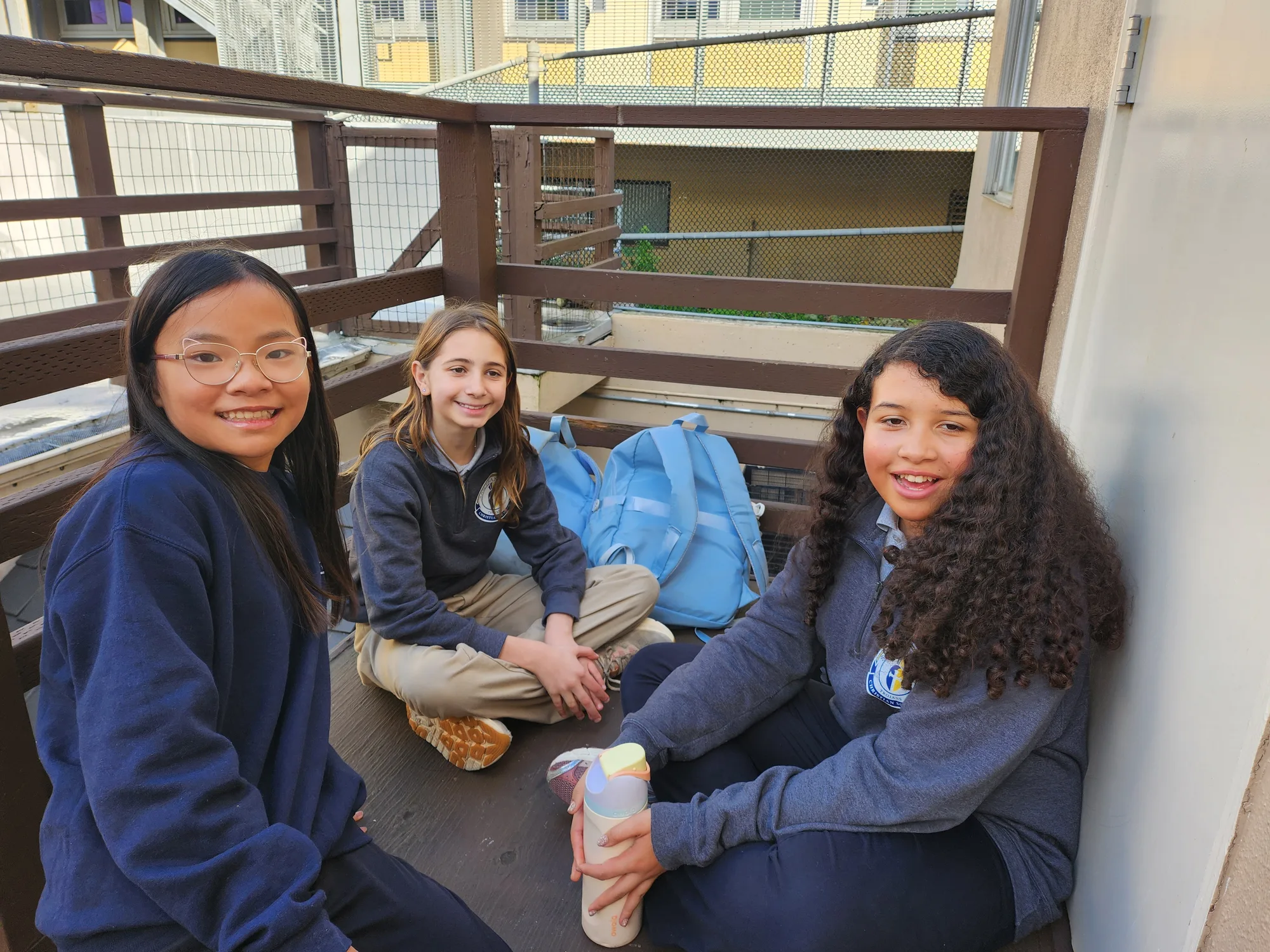 Three fifth-grade girls seated on a deck smiling in navy school quarter-zips