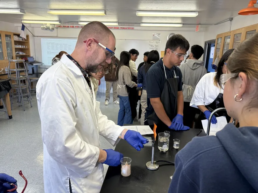 A teacher and high school students in goggles and gloves running a burner experiment in the chemistry lab