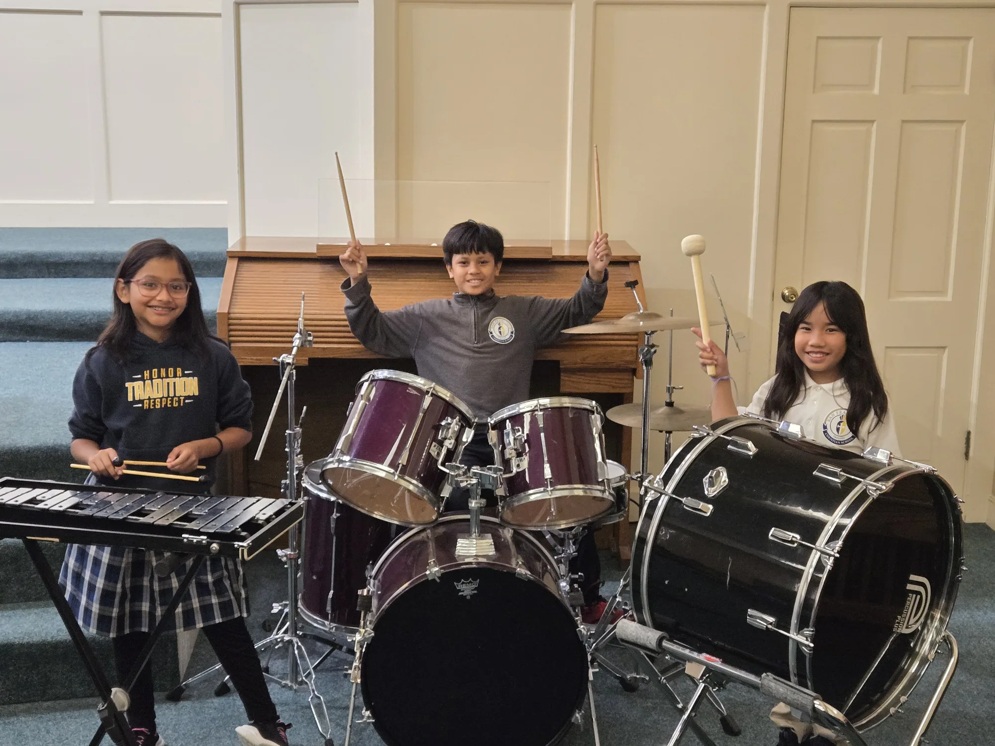 Three students posed at the drum kit with big smiles and raised drumsticks