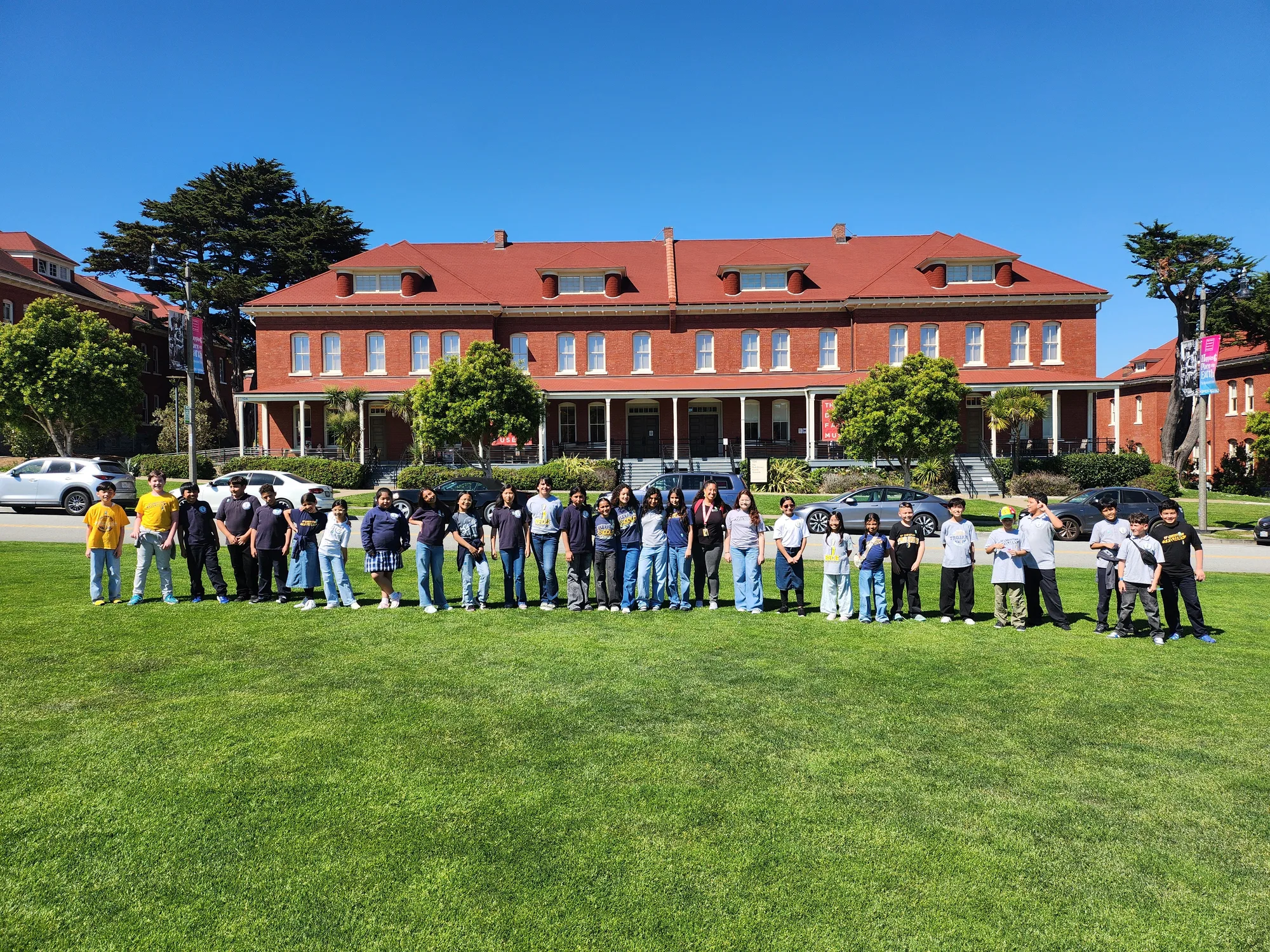 A class posed on the lawn in front of a red Presidio museum building on a sunny day