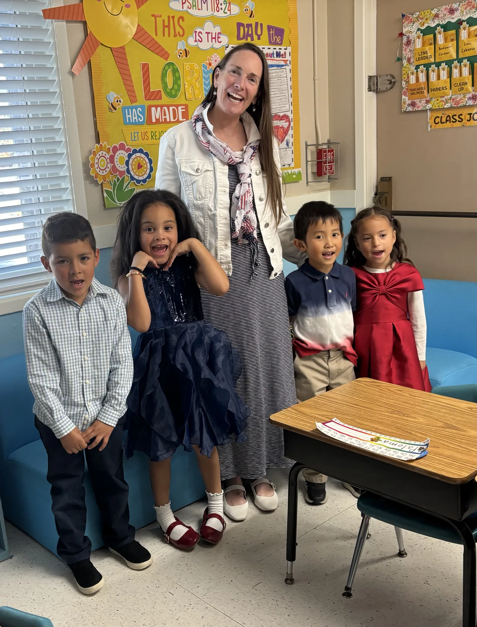 A teacher with four dressed-up kindergartners making joyful faces beneath a Psalm 118:24 classroom board