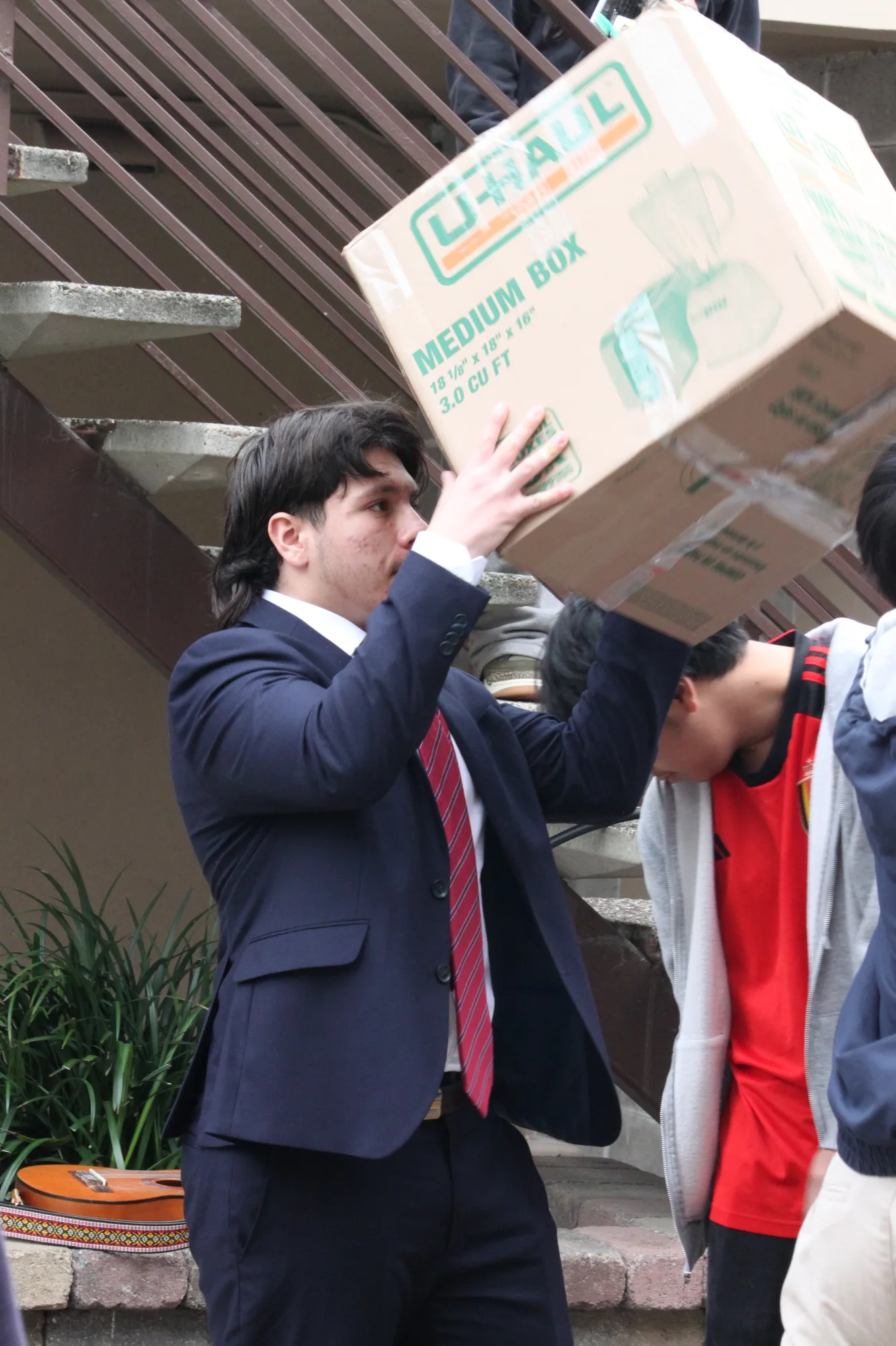 A student in a navy suit hoisting a moving box overhead in a service-day chain
