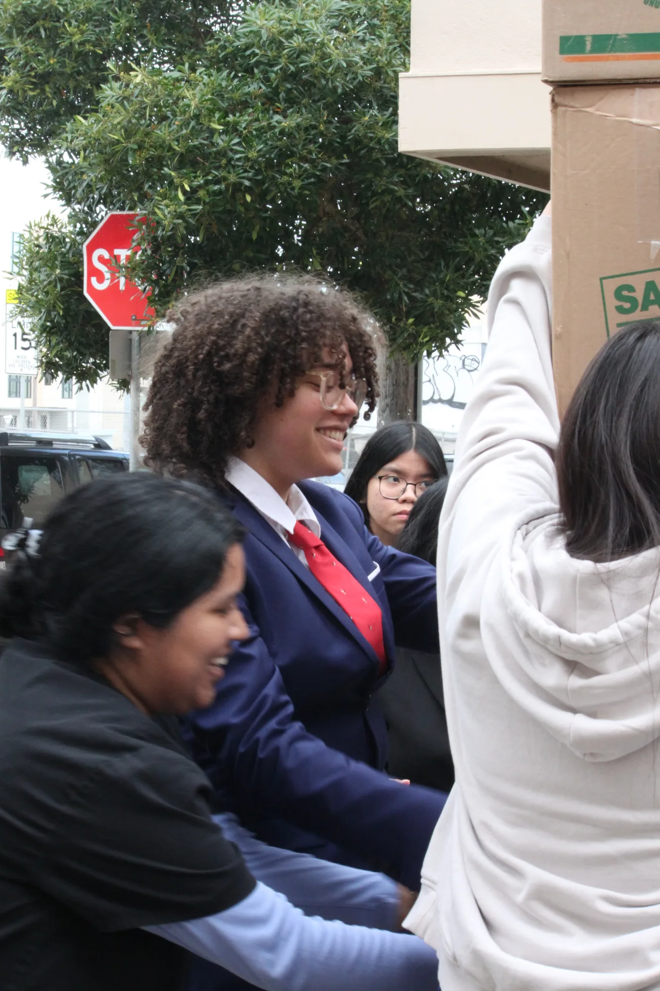 A student in a navy blazer and red tie smiling on a San Francisco street corner